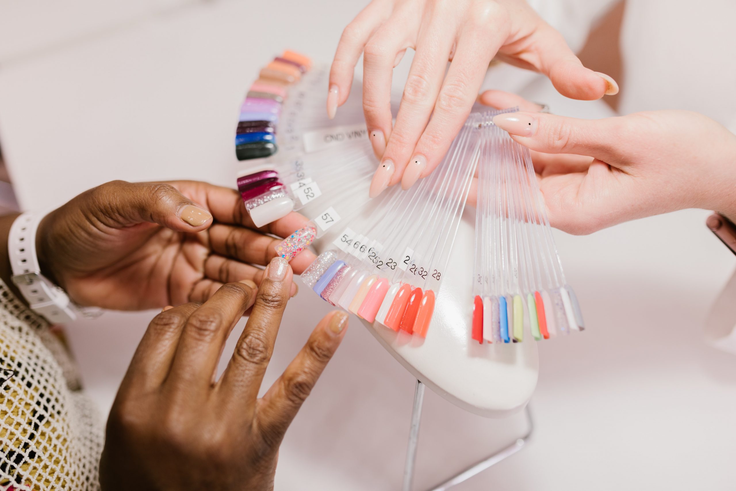 Des mains colorées et soignées, avec des ongles parfaitement vernis dans une teinte pastel. Des produits de beauté sont éparpillés sur une surface en verre, prêts à être utilisés pour une manucure impeccable